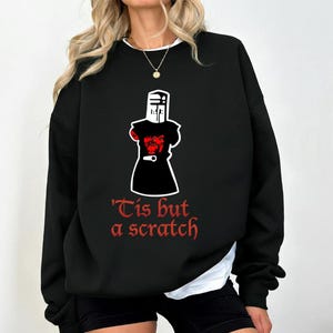 May include: Black sweatshirt featuring a graphic of a knight with the text "'Tis but a scratch" in red. The knight graphic is in white and red. The sweatshirt has long sleeves and a crew neck.