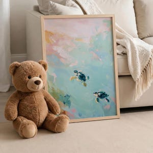 May include: A framed art print featuring an abstract painting of two sea turtles swimming in turquoise water. The artwork has a light wood frame and is displayed next to a brown teddy bear. The painting has pastel colors.