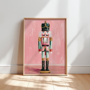 May include: A framed print of a colorful nutcracker soldier. The artwork features a pink background with a painted nutcracker in green, pink, and black. The nutcracker has a white mustache and a black hat with gold accents. The print is in a wooden frame.