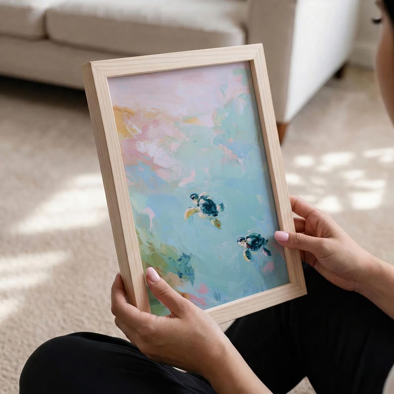 May include: A framed art print featuring an abstract painting of two sea turtles swimming in a turquoise ocean. The artwork has a soft, pastel color palette with pink, blue, and yellow hues. The frame is made of light-colored wood.