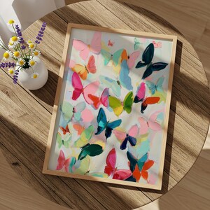 May include: A framed artwork depicting numerous painted butterflies in shades of pink, blue, green, and orange. The butterflies are set against a white backdrop, and the frame is made of light wood. A small vase of flowers is placed beside the artwork.