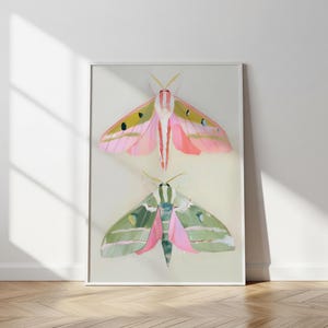 May include: A framed art print featuring two stylized moth illustrations. The top moth is pink with yellow and green accents, while the bottom moth is green and pink. The artwork is set against a neutral background.