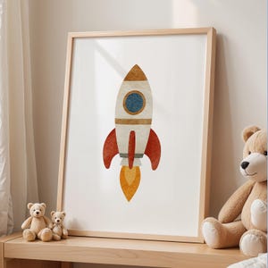 May include: A framed print of a colourful rocket ship illustration. The rocket has a white body with red fins, a blue porthole, and an orange and brown top. A yellow flame bursts from the bottom. The frame is light wood.