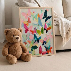 May include: A brown teddy bear sits beside a framed artwork. The artwork depicts numerous colourful butterflies in shades of pink, blue, green, and orange against a light background. The frame is made of light-coloured wood.