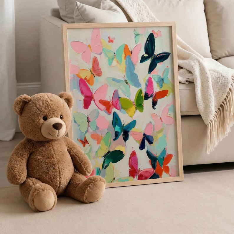 May include: A brown teddy bear sits beside a framed artwork. The artwork depicts numerous colourful butterflies in shades of pink, blue, green, and orange against a light background. The frame is made of light-coloured wood.