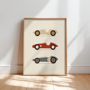 以下が含まれることがあります： クリーム、赤、ブルーグレーの3台のヴィンテージレーシングカーが描かれた額入りプリント。 各車は、黒いホイールと白いインテリアを備えたミニマリストデザインです。 アートワークは、明るい木製のフレームに表示されます。
