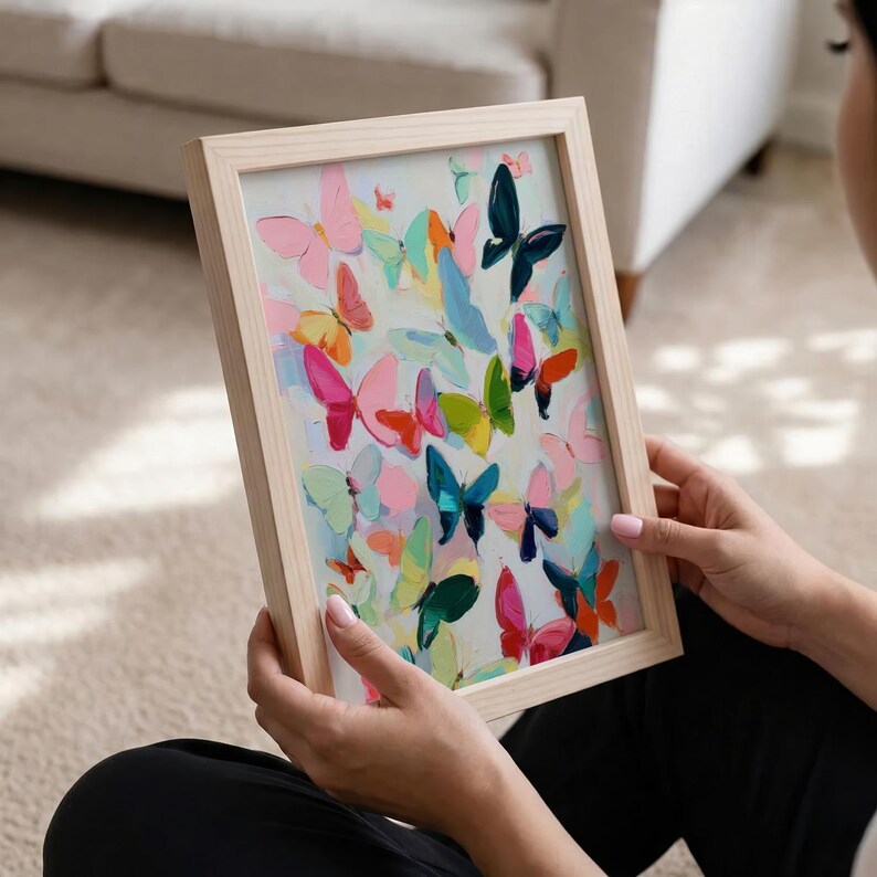 May include: A framed painting with a light wood frame, showcasing a colourful array of butterflies. The artwork features butterflies in pink, blue, green, and orange hues, set against a pale background. The painting is held by a person.