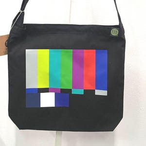 May include: Black tote bag with a colorful test pattern graphic. The bag has a long strap for shoulder or crossbody wear. A small tag is attached to the side. The bag is made of a durable material.