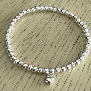 May include: A silver beaded bracelet with a small heart charm. The bracelet is made of small, round, shiny beads strung together. The heart charm hangs from the center of the bracelet. The bracelet is displayed on a light-colored wooden surface.
