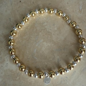 May include: A beaded bracelet featuring alternating gold and silver-colored spheres. The bracelet has a small, round silver charm with the number "925" stamped on it. The bracelet is displayed on a light brown surface.