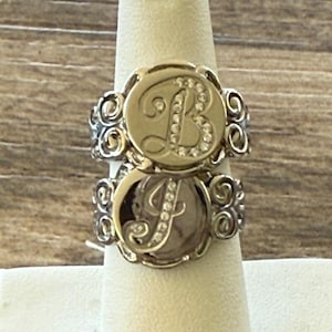 May include: Two stacked silver-toned rings featuring circular faces with ornate, swirling metalwork on the bands. The top ring displays a script letter B, and the bottom ring displays a script letter J, both accented with small, clear embedded stones.