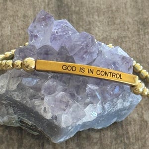 May include: Gold beaded bracelet with a rectangular bar engraved with the words "GOD IS IN CONTROL." The bracelet is displayed on a purple crystal formation. The beads are small and round.