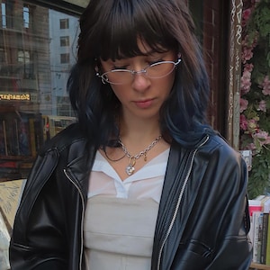 May include: A person wearing a black leather jacket, a white collared shirt, and a beige top. A silver chain necklace with a heart pendant and small charms is visible. The person is also wearing thin-framed glasses.