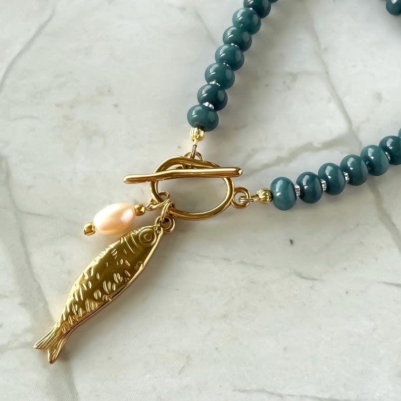 Peut inclure: Un collier de perles bleu-vert avec un pendentif poisson dor&eacute; et un fermoir &agrave; bascule dor&eacute;. Le pendentif comporte un petit charme de perle rose clair. Le collier est sur une surface en marbre blanc et gris.
