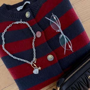 May include: A navy blue and red striped cardigan with round buttons, a silver chain necklace with charms, clear-framed glasses, and a black purse. The cardigan is folded, and the items are arranged on a light-colored surface.
