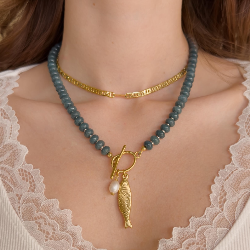 Peut inclure: Un collier superpos&eacute; avec une cha&icirc;ne dor&eacute;e et un collier de perles avec un pendentif poisson dor&eacute; et une perle. Le collier de perles est de couleur bleu-vert fonc&eacute;. Le collier est port&eacute; autour du cou.
