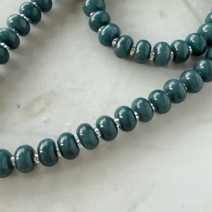 Peut inclure: Gros plan sur un collier de perles de couleur teal. Les perles ovales sont enfil&eacute;es avec des entretoises argent&eacute;es. Le collier est pr&eacute;sent&eacute; sur une surface en marbre blanc.