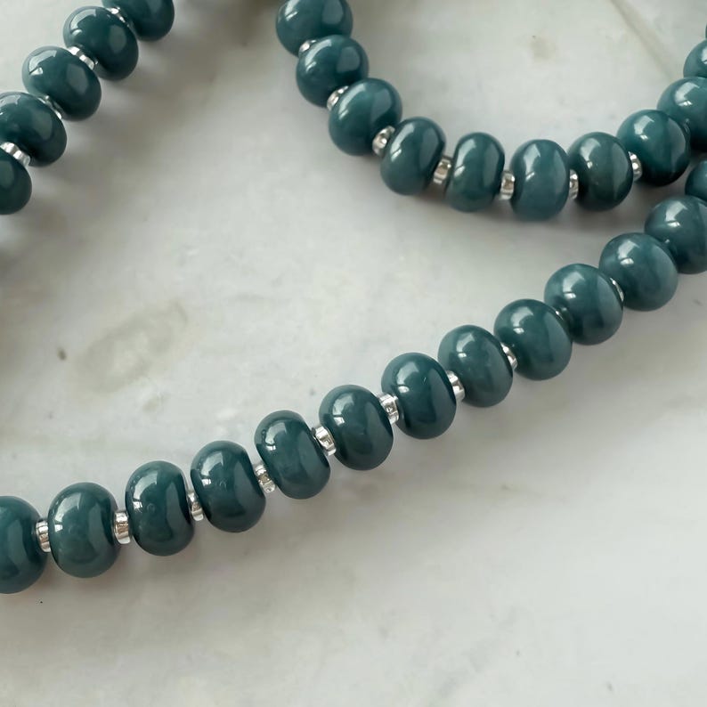 Peut inclure: Gros plan sur un collier de perles de couleur teal. Les perles ovales sont enfil&eacute;es avec des entretoises argent&eacute;es. Le collier est pr&eacute;sent&eacute; sur une surface en marbre blanc.