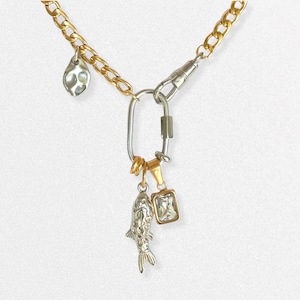 May include: A gold and silver-toned necklace with multiple charms. The necklace features a gold chain, a silver carabiner clasp, and various charms, including a fish, a rectangular gem, and a textured teardrop. The necklace is displayed against a white background.