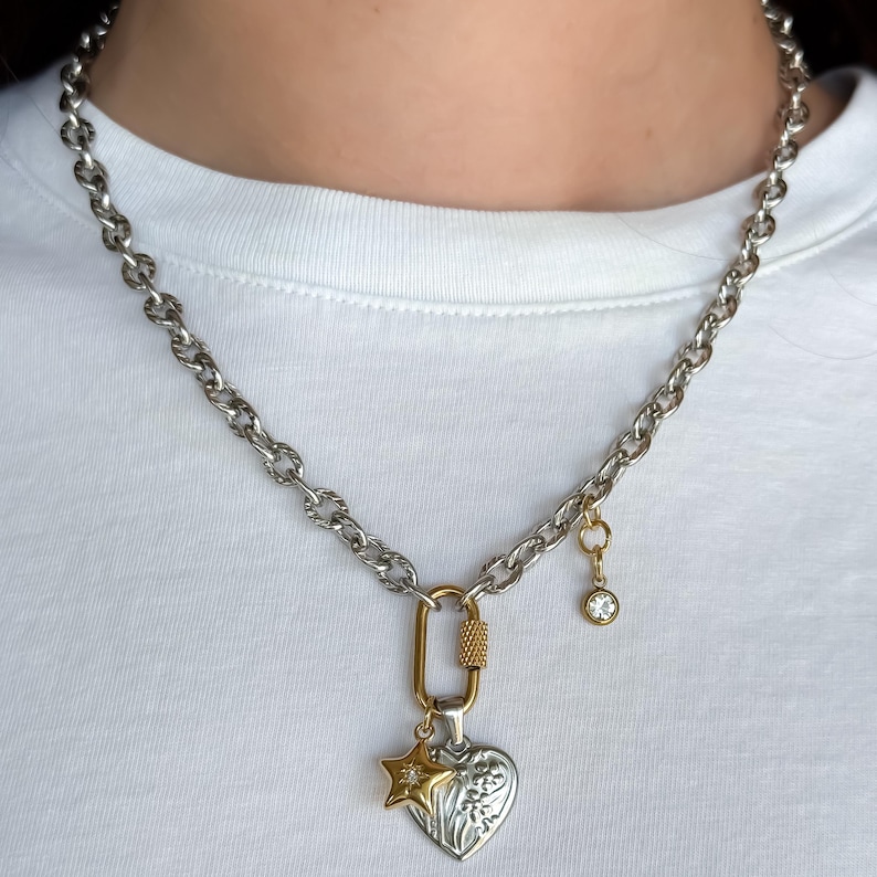 May include: Silver-tone chain necklace with a gold-tone clasp and charms. The charms include a heart pendant with floral details, a star, and a small dangling charm with a clear crystal. The necklace is worn against a white background.