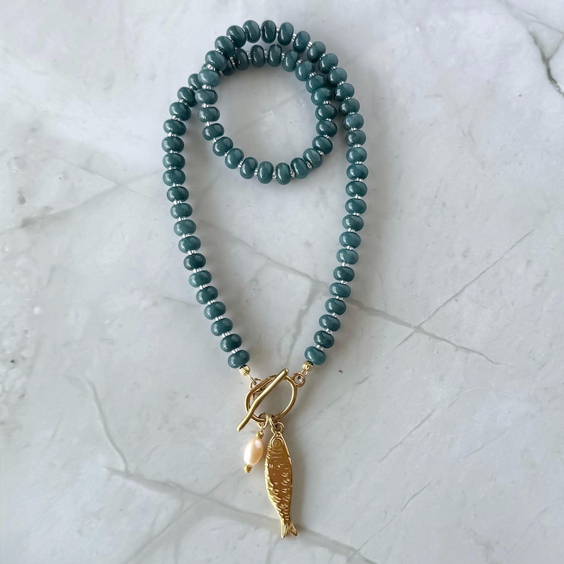 Peut inclure: Collier de perles turquoise avec fermoir dor&eacute; et pendentif poisson. Le collier est dot&eacute; d'un fermoir &agrave; bascule et d'une petite perle rose clair. Les perles sont d'un bleu-vert profond.
