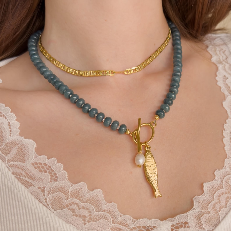 Peut inclure: Ensemble de deux colliers. La cha&icirc;ne sup&eacute;rieure est en or. Le collier inf&eacute;rieur est un collier de perles avec un pendentif poisson dor&eacute; et une perle. Les perles sont de couleur bleu-vert. Le pendentif poisson est dor&eacute;.