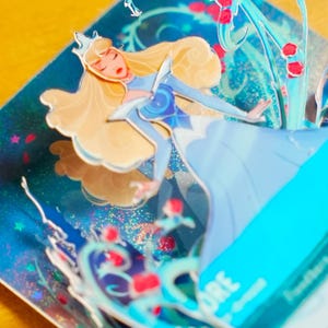 May include: Close-up of a 3D pop-up card featuring Aurora, the Disney princess, in a light blue dress. The card has a sparkly, iridescent background with glitter and images of roses and vines.  Aurora's blonde hair and crown are clearly visible. The card also includes text that says 'AURORA'.
