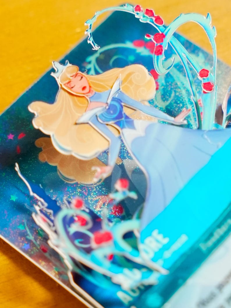 May include: Close-up of a 3D pop-up card featuring Aurora, the Disney princess, in a light blue dress. The card has a sparkly, iridescent background with glitter and images of roses and vines.  Aurora's blonde hair and crown are clearly visible. The card also includes text that says 'AURORA'.