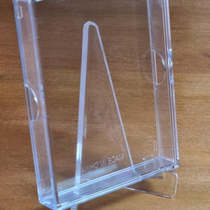 May include: Clear plastic display case with a built-in easel stand. The case is designed to hold and display items such as photos or small collectibles.  Made in China is printed on the base. The transparent design allows for visibility of the contents.  The stand provides a stable upright display position.  Suitable for various display purposes.