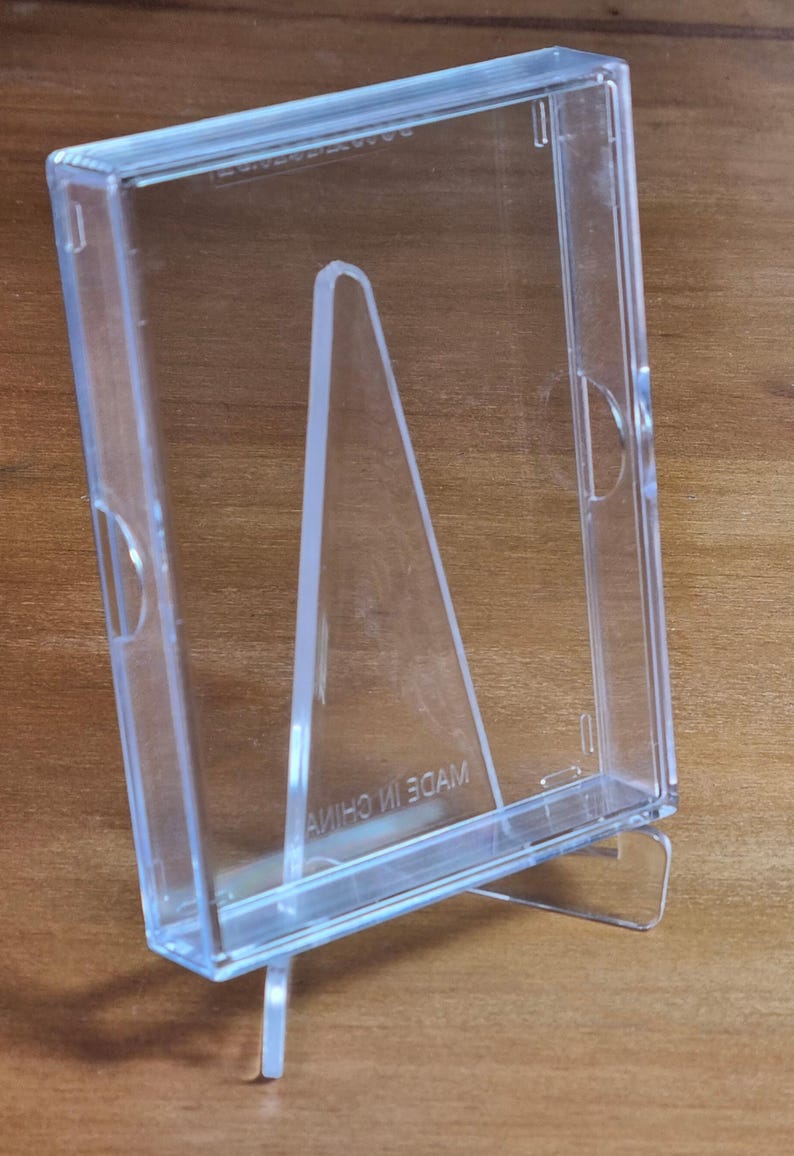 May include: Clear plastic display case with a built-in easel stand. The case is designed to hold and display items such as photos or small collectibles.  Made in China is printed on the base. The transparent design allows for visibility of the contents.  The stand provides a stable upright display position.  Suitable for various display purposes.