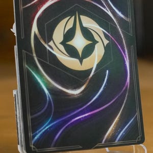 May include: Close-up view of a card with a dark background featuring swirling, multicolored neon lines. A central, light beige hexagonal emblem with a glowing, four-pointed star is prominently displayed. The card is displayed on a clear acrylic stand. The overall aesthetic suggests a fantasy or mystical theme.