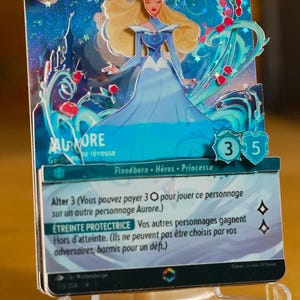 May include: A collectible card game featuring Aurora, a princess. The card is predominantly teal and blue, with a 3D sculpted image of Aurora in a flowing gown.  The card's text includes 'Alter 3', 'Etreinte Protectrice', and game stats.  The card's background includes stars and floral elements.  The card is displayed on a clear acrylic stand.