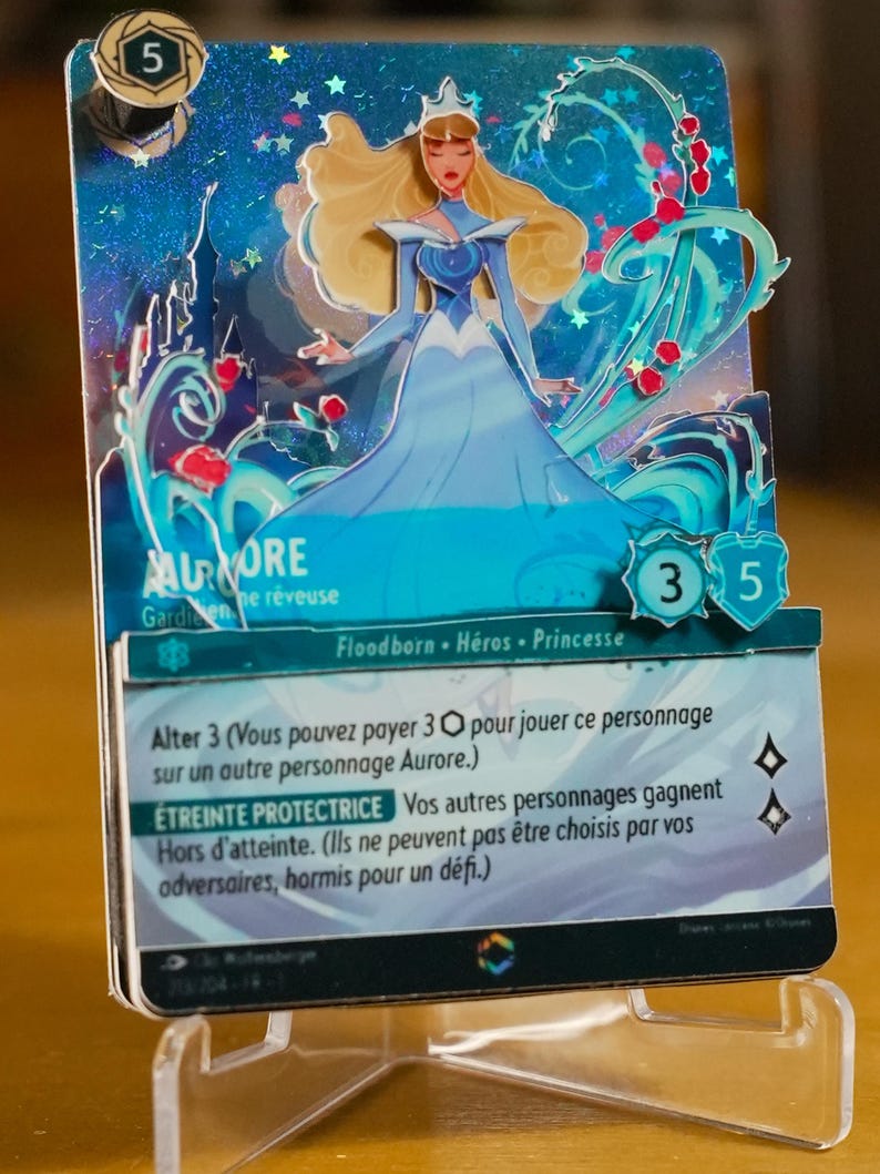 May include: A collectible card game featuring Aurora, a princess. The card is predominantly teal and blue, with a 3D sculpted image of Aurora in a flowing gown.  The card's text includes 'Alter 3', 'Etreinte Protectrice', and game stats.  The card's background includes stars and floral elements.  The card is displayed on a clear acrylic stand.