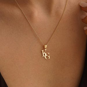 Puede incluir: Un collar de tono dorado con un colgante que presenta las letras K y G en una fuente con serifa. El colgante cuelga de una delicada cadena de tono dorado. Las letras están colocadas verticalmente con la K sobre la G, creando un dije inicial.