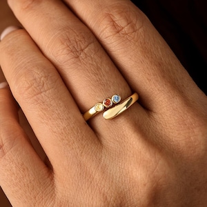 May include: A gold bypass ring featuring three small, round, bezel-set gemstones in yellow, orange, and blue. The band has a smooth, polished finish and an open design that overlaps slightly at the centre, showcasing the three colourful stones.