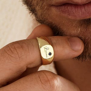 May include: A gold signet ring featuring a smooth, polished surface with the name Lucas engraved in a cursive script, accompanied by a small, dark, star-shaped accent stone set directly next to the lettering on the face of the ring.