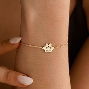 May include: A delicate gold bracelet featuring a paw print charm with the name "Indie" engraved on it. The bracelet is worn on a wrist, showcasing its fine chain and detailed charm. The jewelry piece is a personalized accessory.