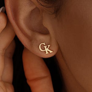 May include: Gold-colored stud earrings with the initials "GK" are displayed on an ear. The earrings are small and simple, with a classic design. The image is a close-up, highlighting the jewelry's details.