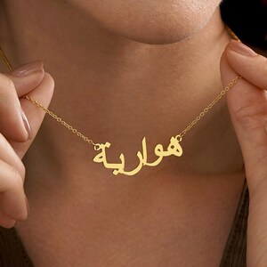 May include: A gold-colored necklace with Arabic script spelling a name. The necklace is held by a person, showcasing the delicate chain and the personalized pendant. The pendant is in a cursive font.