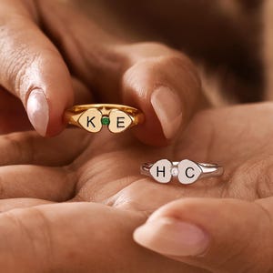 Peut inclure: Une bague dorée avec deux cœurs gravés des lettres K et E, séparés par une petite pierre verte, et une bague argentée avec deux cœurs gravés des lettres H et C, séparés par une petite pierre transparente au centre de chaque bijou.