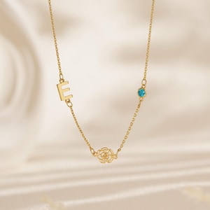 Pode incluir: Um delicado colar dourado com a letra "E", uma pequena pedra preciosa turquesa e um pingente em forma de rosa. O colar é exibido em um fundo macio e esbranquiçado, destacando seu design elegante. O colar é da Antique Trinket Co.