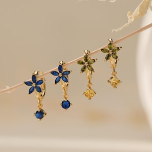 May include: Four gold hoop earrings featuring flower-shaped charms with marquise-cut gemstones and a single dangling round gemstone. Two earrings have blue stones, and two have olive green stones with yellow dangling accents.