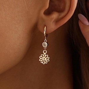 May include: A gold hoop earring featuring a small, clear, round gemstone charm suspended above a gold flower-shaped pendant. The earring is secured through the earlobe, showcasing a delicate design with a polished metallic finish and a clear stone.