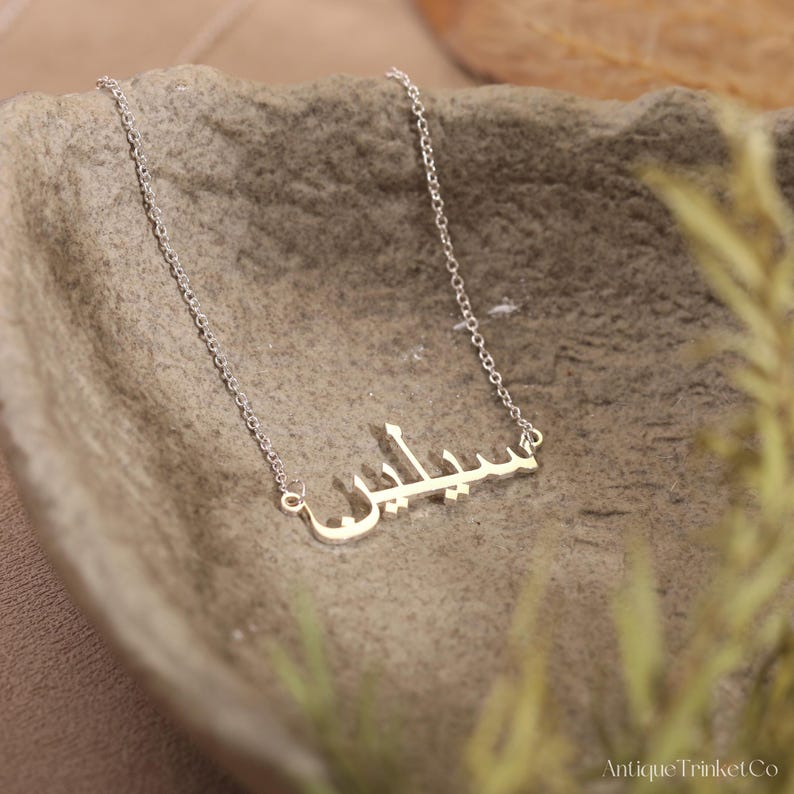 May include: A silver-colored necklace with an Arabic name pendant. The pendant is in a script font and is suspended from a delicate chain. The necklace is displayed on a textured, gray surface.