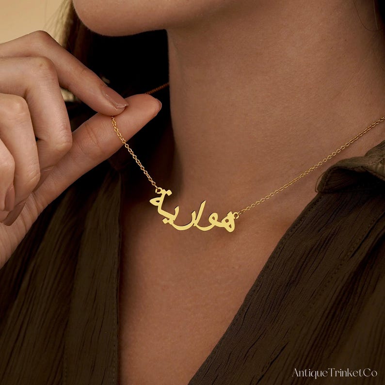 May include: A gold-colored necklace with an Arabic name pendant. The pendant features the name "Hawa" in elegant script. The necklace is displayed against a neutral background, highlighting the jewelry's design and color.