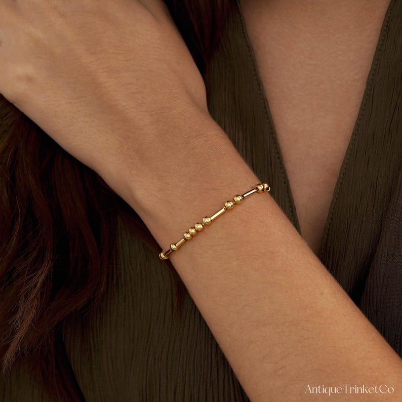 May include: A gold beaded bracelet with a mix of small round beads and cylindrical bars. The bracelet is worn on a person's wrist, against a dark green background. The bracelet is a delicate piece of jewelry.