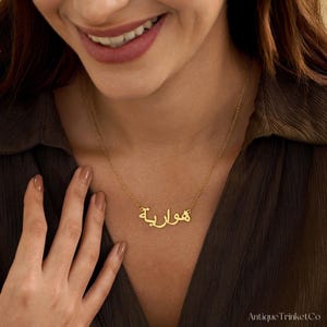May include: A gold-colored necklace with the Arabic name "هوارية" displayed in elegant script. The necklace is on a delicate chain. The image also shows the AntiqueTrinket Co. logo.