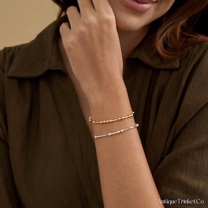 May include: Two beaded bracelets, one gold and one silver, are worn on a person's wrist. The gold bracelet is on top, and the silver bracelet is below. The bracelets have small, round beads and are simple in design.