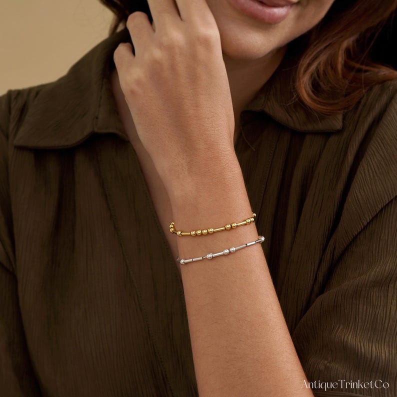 May include: Two beaded bracelets, one gold and one silver, are worn on a person's wrist. The gold bracelet is on top, and the silver bracelet is below. The bracelets have small, round beads and are simple in design.