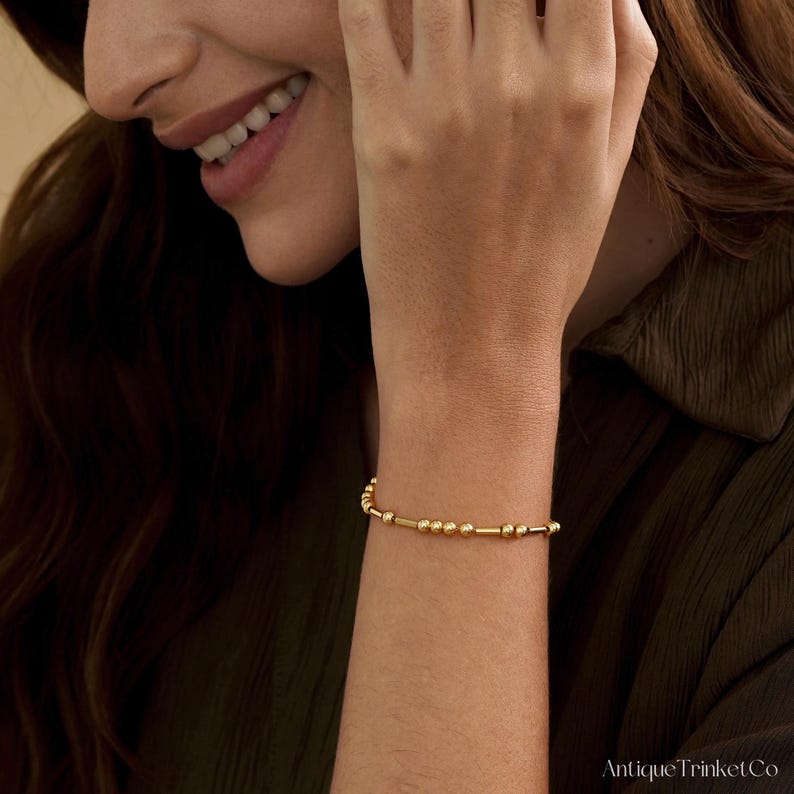 May include: A gold beaded bracelet with a delicate design. The bracelet features small, round beads interspersed with longer, cylindrical gold bars. The jewelry is worn on a wrist, showcasing its simple elegance.